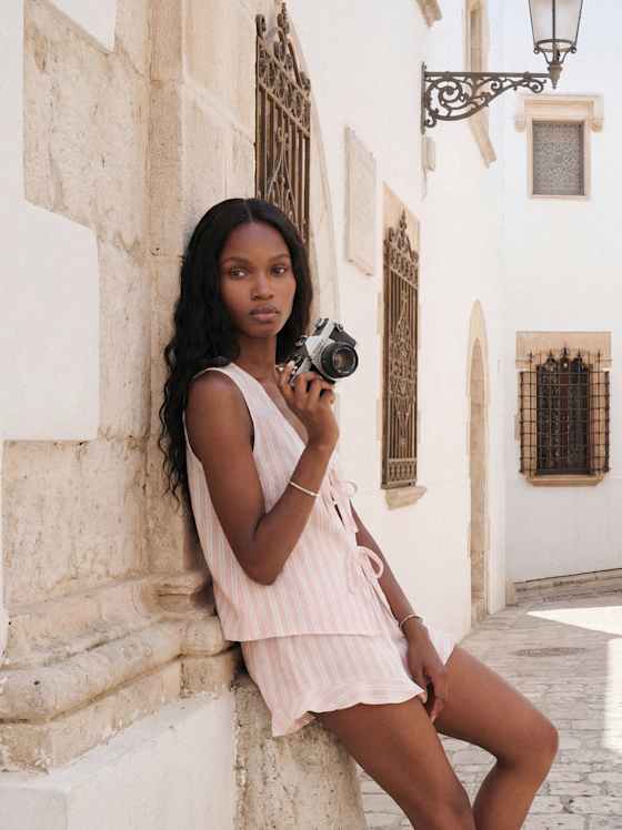 Person in roséfarbenem, gestreiftem Sommerset mit Shorts lehnt lässig an Sandsteinmauer und hält eine Kamera, sonnige helle Gasse im Hintergrund.