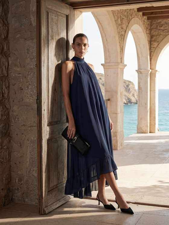 Person im fließenden, dunkelblauen Highneck-Kleid mit schwarzer Clutch und Slingpumps lehnt an offener Tür, im Hintergrund Arkaden und Meer.
