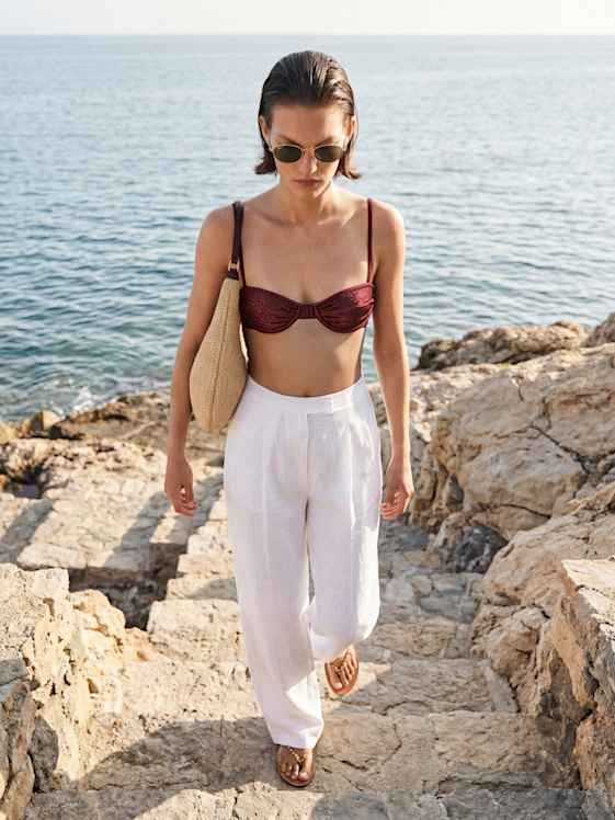 Person im bordeauxroten Bikini-Oberteil und weißen Leinenhosen geht eine steinerne Treppe am Meer hinauf, Strohtasche über der Schulter, sonnige Küste