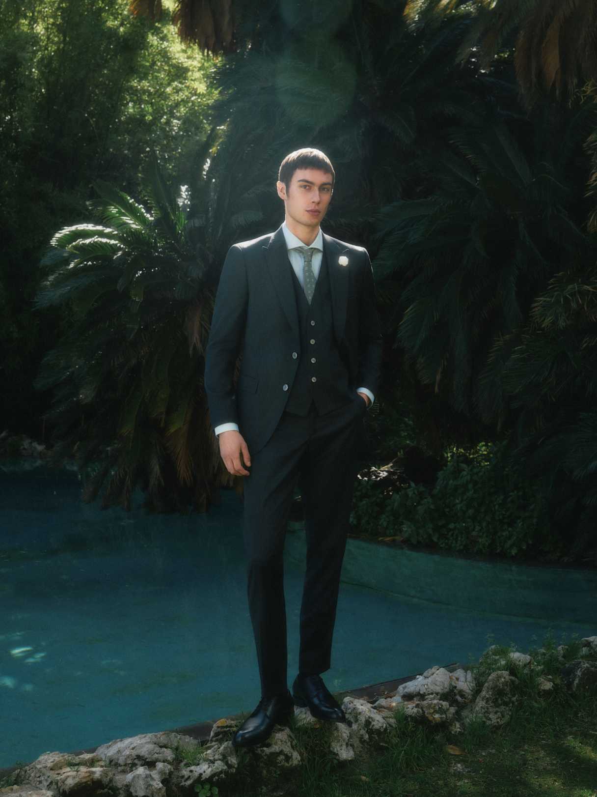 Elegant gekleidete Person im dunklen Anzug steht am Rand eines Pools im Garten, umgeben von dichtem Grün und sanftem Licht.