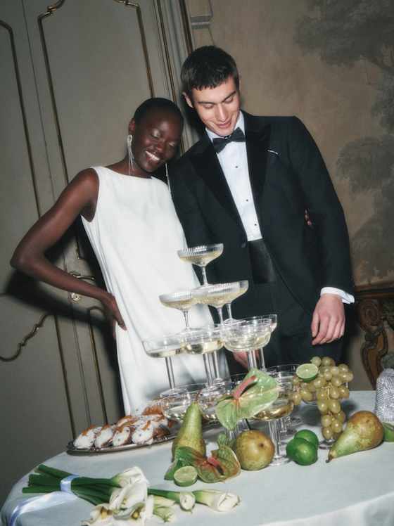 Zwei elegant gekleidete Personen stehen an einem festlich gedeckten Desserttisch mit Etagere, Gebäck und Früchten bei einer Feier.