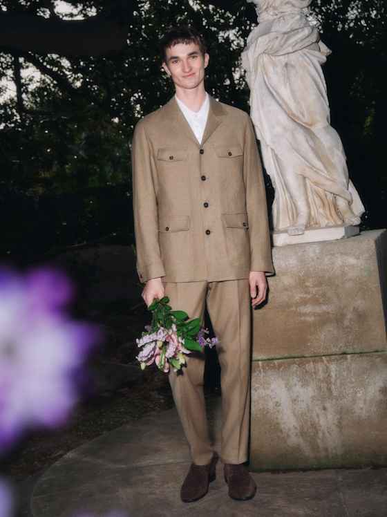 Elegant gekleidete Person in beigem Anzug steht im Garten neben einer Statue und hält einen kleinen Blumenstrauß in der Hand.