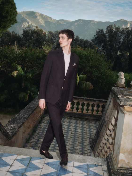 Elegant gekleidete Person im dunklen Anzug geht über eine Terrasse mit Mosaikboden, vor üppigem Grün und Berglandschaft im Hintergrund.