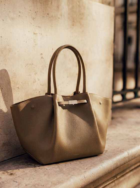 Beigefarbene, strukturierte Ledertasche mit zwei Henkeln, stehend auf einem Steinpodest in der Sonne, unscharfer Hintergrund mit Geländer.