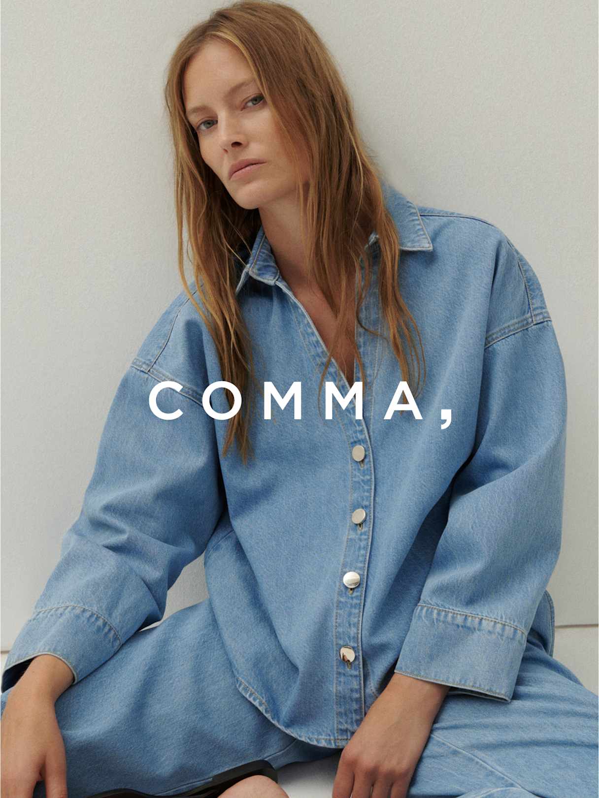Person in weiter Jeansbluse und passender Hose vor hellem Hintergrund, „Comma,“ im Vordergrund.