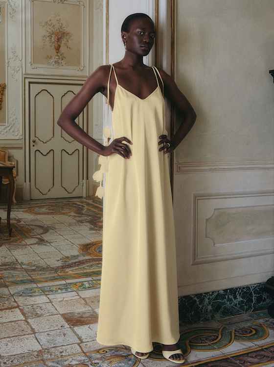 Eine Person steht in einem eleganten, historischen Raum und trägt ein bodenlanges, fließendes Kleid in hellem Gelb mit schmalen Trägern.