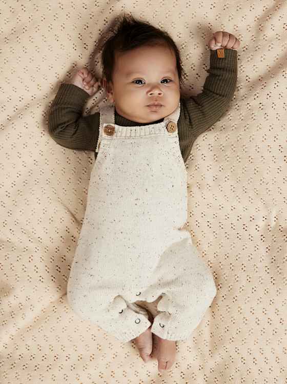 Baby liegt auf heller Decke, trägt einen gemusterten Strampler in Creme mit langärmligem Oberteil in Olivgrün.
