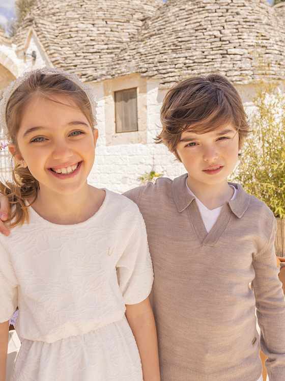 Zwei Kinder tragen hochwertige cremefarbige Outfits, ein Kleid und ein Pullover mit V-Ausschnitt