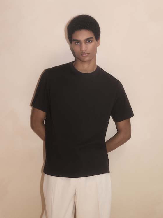 Schwarzes Kurzarm‑T‑Shirt, kombiniert mit heller Stoffhose vor neutralem Hintergrund.