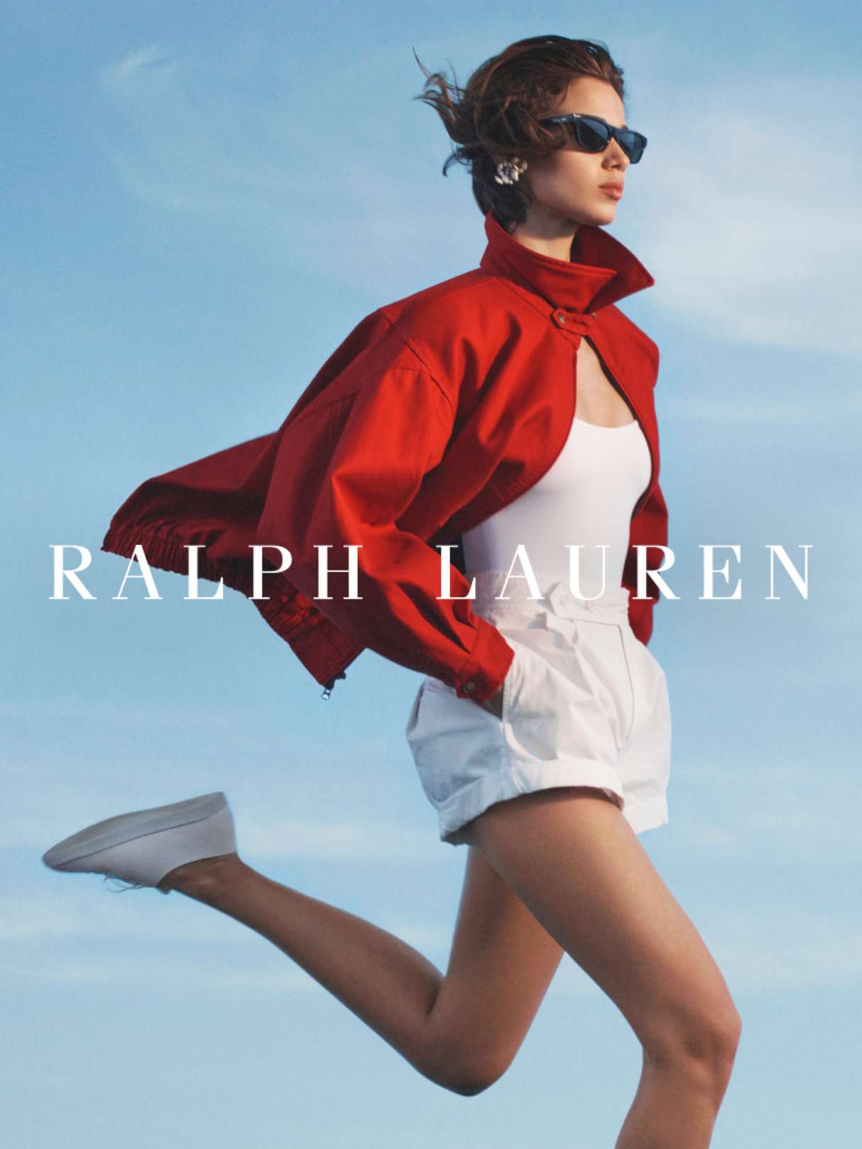 Person in roter Jacke, weißer Shorts und weißen Sneakern springt vor blauem Himmel, Markenlogo „Ralph Lauren“ im Vordergrund.