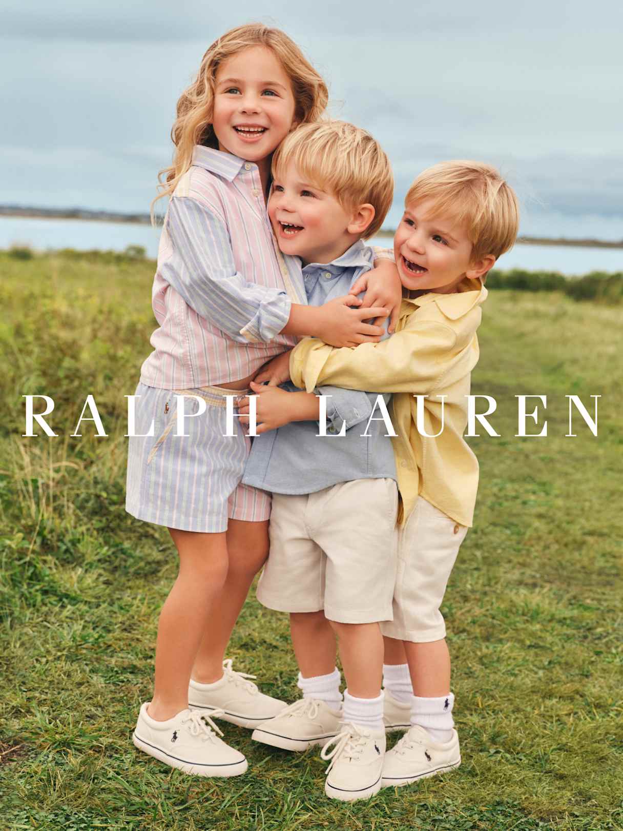 Drei Kinder in sommerlicher Ralph-Lauren-Kleidung umarmen sich auf einer Wiese, „Ralph Lauren“ im Vordergrund.